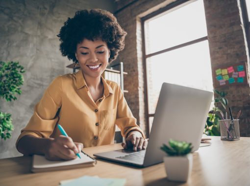 A young professional writing on a notepad at a table with a laptop in front of her.